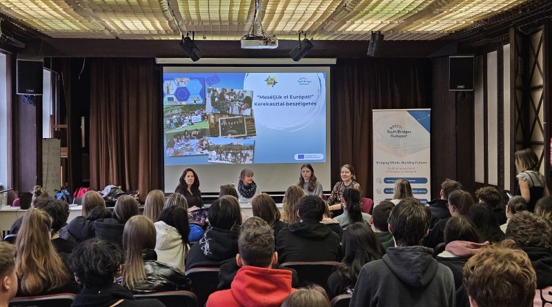 Roundtable discussion of 4 women in a high school with students in the audience.